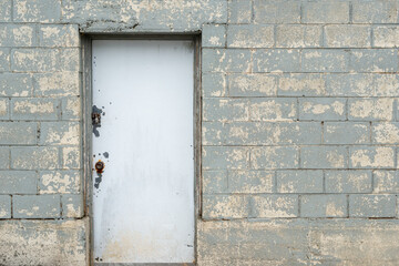 An exterior industrial solid white metal door with a lock and handle in an old grey concrete block wall in a building. The gray blocks' paint is worn and scuffed in places.  