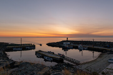 A small fishing village and marina at sunset. The ocean is smooth and reflecting the dramatic orange sky. There's a breakwater dividing a small cove as a recreational fishing boat enters the harbour.