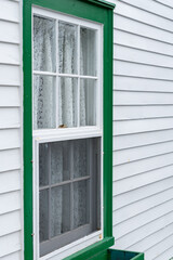 Obraz premium The exterior outside wall of a stark white heritage wood building with vibrant green trim. The country style house has narrow clapboard and a single double hung window with lace curtains hanging. 