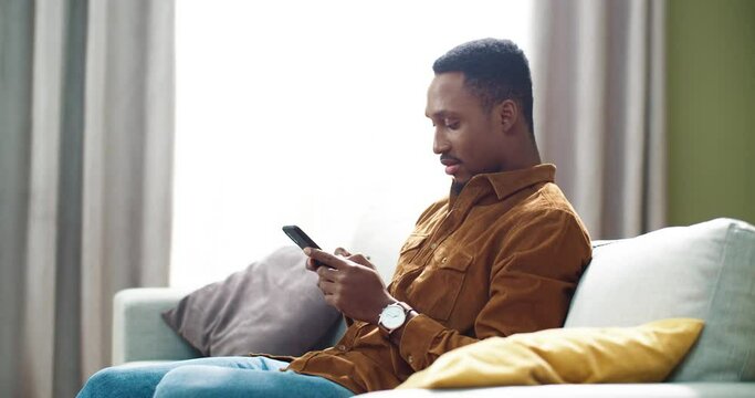 Young handsome african american man sitting at home on sofa with phone in hands and typing messages to friends.
