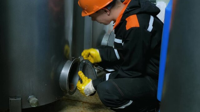 A worker wearing a helmet and gloves, removes the hatch cover to replace the filter element in the water filtration tank. Industrial profession. Dangerous job