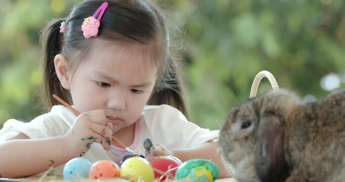 Close up of asian little girl coloring easter eggs with colors and brush near a brown rabbit on easter festive desk. Preparation of the Easter. Zoom in, slow motion