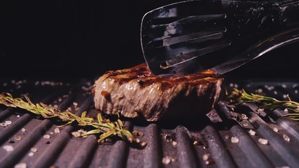 Delicious juicy meat steak cooking on grill. Aged prime rare roast grilling marble beef. Electric roaster, rosemary black pepper, salt. Kitchen tongs.