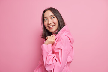 Pretty glad brunette Asian woman stands half turned against pink studio background has good mood...