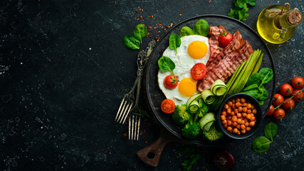 English breakfast: chickpeas, avocados, scrambled eggs, bacon vegetables on a black stone plate. Top view. Free space for text.