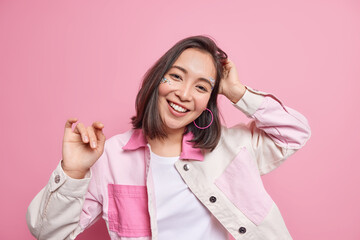 Horizontal shot of optimistic carefree Asian woman tilts head smiles sincerely dressed in fashionable jacket being in good mood isolated over pink background. Happy emotions and feelings concept