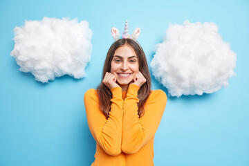 Attractive young woman with dark long hair keeps hands under chin smiles gently wears unicorn headband and orange jumper isolated over blue background white clouds above. Positive emotions concept