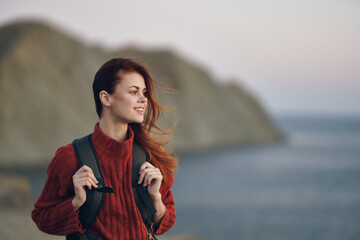 woman hiker holds a backpack on her shoulders and look at the sea from the side of the mountains in the background