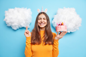 Birthday celebration concept. Positive brunette European woman crosses fingers makes wish befor burning candles on festive cake smiles broadly isolated over blue background with white clouds.