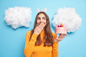 Happy dreamy brunette woman celebrates birthday holds strawberry cake covers mouth and chuckles dressed in casual jumper isolated over blue background with white clouds above. Festive occasion