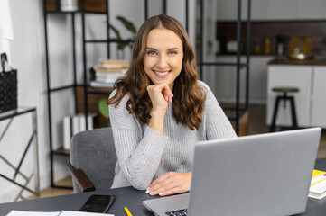 Fototapeta na wymiar Executive female employee in casual wear sitting at the office desk in front of the laptop, friendly woman looks at the camera and smiles, balanced and tranquility freelancer woman on the workplace