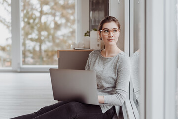 Young woman sitting working on a laptop on the floor leaning on a window