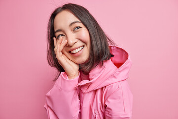 Happy brunette millennial female model looks with sincere cheerful expression keeps hand on face...