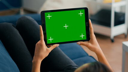 Close up of woman holding in horizontal mode tablet computer with mock up green screen chroma key display lying on sofa. Freelancer using isolated touchscreen device for social networks browsing