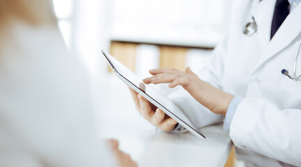 Unknown male doctor and patient woman discussing current health examination while sitting in clinic and using tablet computer. Perfect medical service in hospital. Medicine and healthcare concept