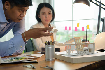 Asian architect-designer holding the part of the model of modern box house while discussing with team people about concept of building and construction projects. Focusing on his hand holding model. 