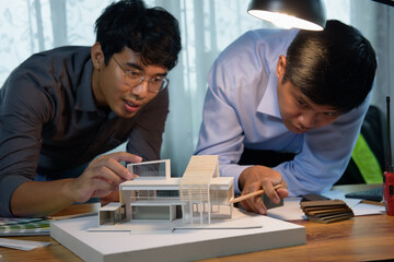 Asian architects discussing with designer or civil engineer about concept of building design and construction project for modern box house. Focusing on the house model.