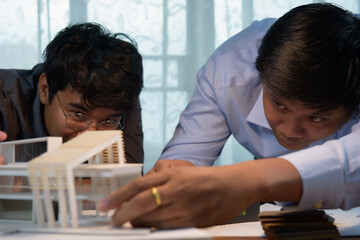 Asian architects discussing with designer or civil engineer about concept of building design and construction project for modern box house. Focusing on the house model.