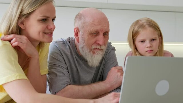 Young children teach an elderly grandfather to work at modern notebook gadget at home. Elderly retired grandfather in circle of his family daughter and granddaughter is trained work on modern laptop.