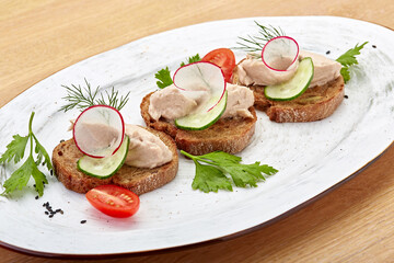 Bruschetta with pate, cucumber and radish, garnished with herbs and tomatoes on a beautiful white oval plate. View from above. 