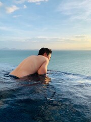 Young man in outdoor infinity swimming pool seeing beautiful ocean and sky view during sunset time Travel lifestyle and leisure concept