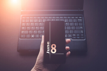 June 8th. Day 8 of month, Calendar date. Hand Holding Mobile Phone on Laptop Computer dark background with sunshine. Summer month, day of the year concept.