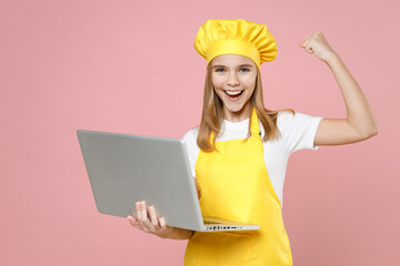 Teen girl chef cook confectioner housewife baker in yellow apron white t-shirt cap hold laptop pc computer do winner gesture clench fist isolated on pink background studio portrait Food cake concept. - obrazy, fototapety, plakaty