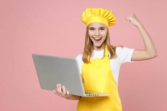 Teen girl chef cook confectioner housewife baker in yellow apron white t-shirt cap hold laptop pc computer do winner gesture clench fist isolated on pink background studio portrait Food cake concept.