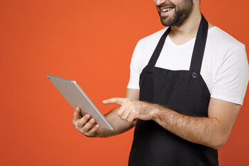 Close up cropped photo portrait shot young man barista bartender barman employee in black apron white t-shirt work in coffee shop use tablet pc computer isolated on orange background business startup