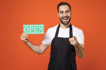 Young man barista bartender barman employee in black apron white tshirt work in coffee shop hold card sign OPEN title text point finger camera you isolated on orange background Small business startup