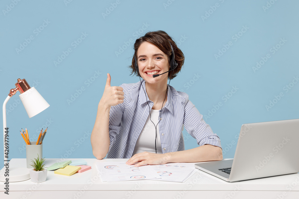 Wall mural Young operator employee business woman in shirt set microphone headset for helpline assistance sit work at office desk with pc laptop show thumb up gesture isolated on pastel blue background studio.