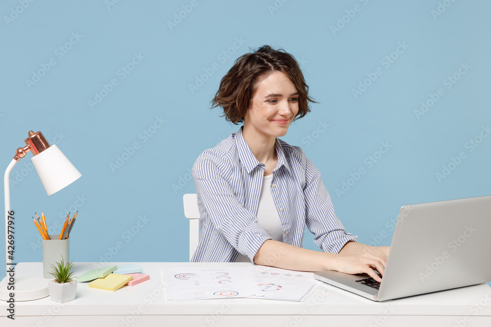 Wall mural Young successful secretary employee business woman wear casual shirt sit work at white office desk with pc laptop computer browsing internet online isolated on pastel blue background studio portrait.