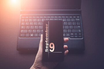 September 9th. Day 9 of month, Calendar date. Hand Holding Mobile Phone on Laptop Computer dark...
