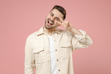 Young smiling happy positive fun fashionable caucasian man in jacket white t-shirt cover eye with victory v-sign gesture isolated on pastel pink background studio portrait People lifestyle concept
