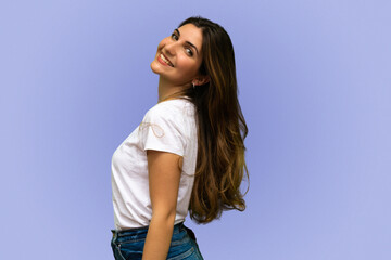Close-up profile portrait of smiling cute young brunette girl with long haircut, wearing white t-shirt with blue jeans, facing left side of blank copy, standing optimistic lilac background © nikcattuto