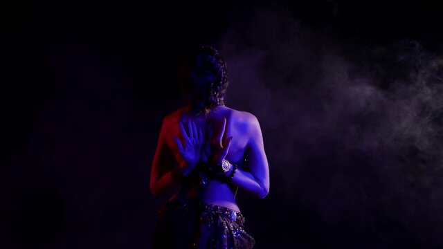 A woman, seen from the back, dancing an oriental dance in a smoky studio on a black background, makes beautiful movements with her hands, she is dressed in a chic suit and illuminated by neon light.