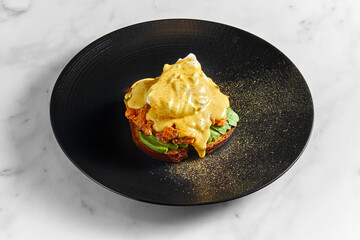 Appetizing and healthy breakfast - toast with avocado, baked tomatoes, poached egg and hollandaise sauce, served in a black plate on a marble background