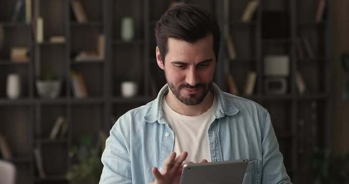 Happy young dreamy sincere man reading message or email with pleasant news on digital computer tablet, looking in distance, visualizing or planning future events, feeling excited alone at home.
