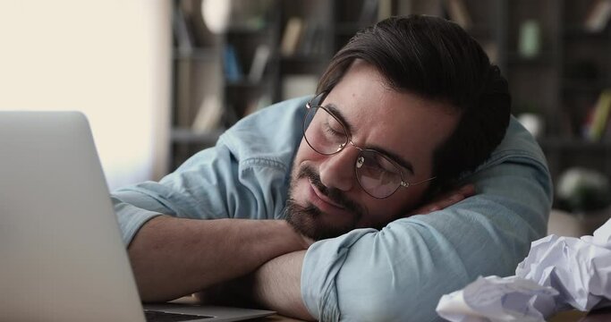 Head shot lazy unproductive tired young male office worker employee in glasses sleeping at workplace on table with laptop and crumpled paper documents, feeling exhausted after overtime workday.