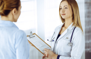 Young woman-doctor and her patient are discussing patient's current health examination, while...