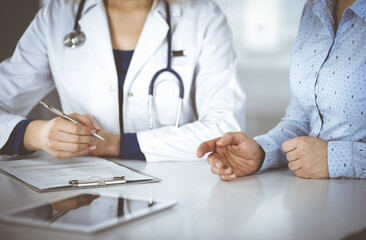 Unknown woman- doctor is listening to her patient, while sitting together at the desk in the cabinet in a clinic. Female physician with a stethoscope is writing at clipboard, close up. Perfect medical