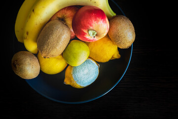 A dark blue plate with ripe colorful whole fruits: green and red apples, kiwi, yellow lemons, bananas. Concept spoiled lemon with white light textured mold. Top view