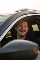 Man sitting in the seat of a new car, smiling while looking out of the window, wearing dark clothes. Caucasian man with dark hair and a short beard. In the evening in a dark grey car.