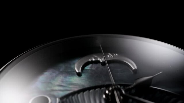 Silver second arrow running on nacrous clockface of rotating swiss made watch with smiling moon. Closeup of metallic details and ribbed pattern on black background