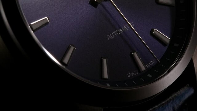 Silver second arrow running on dark blue clockface through the automatic lettering. Closeup of swiss watch with leather strap and grey metallic body. Zoom-out effect