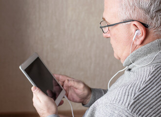 An elderly man with glasses and headphones, with a tablet in his hand, is studying online courses. Distance learning concept
