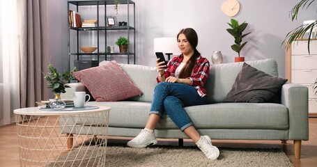 Obraz premium Brunette woman checking credit card balance and celebrating success using smartphone. Female customer excited by money refund or shopping offer, while reading news