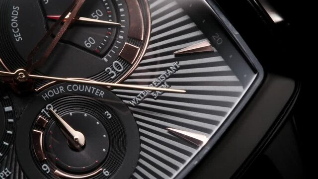 Water resistant swiss watch with black square body, chonograph, stopwatch and hour counter. Golden second arrow running on striped clockface. Extreme closeup