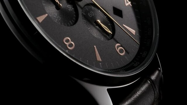 Golden second arrow running on the black clockface of luxury swiss watch with chronograph and leather strap. Closeup with zoom-in effect on black background