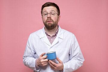 Tired Caucasian doctor holds a mug of coffee in hands, he falls asleep and tries to cheer up. Pink background.